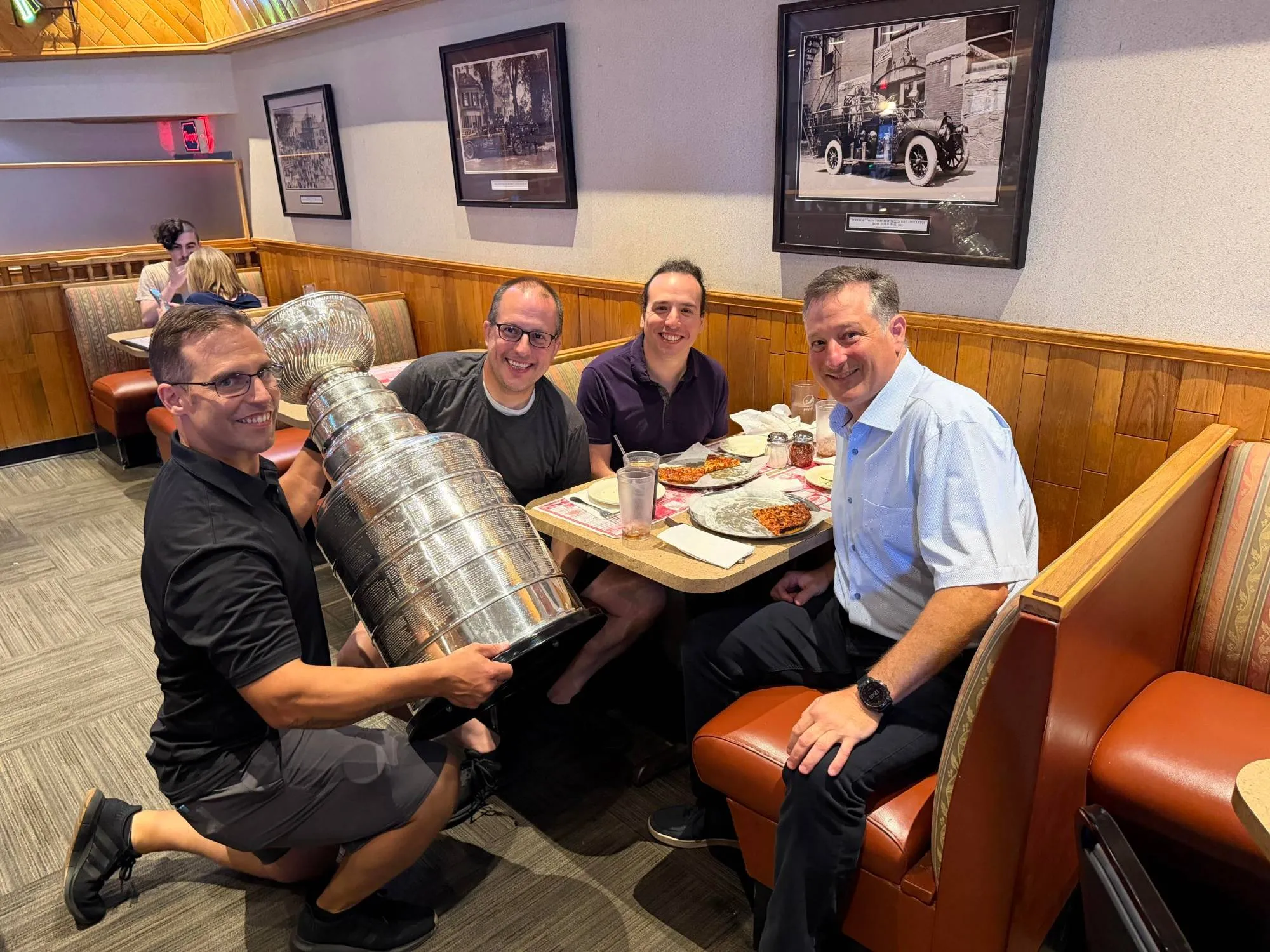 Four members of the Resilient Link team at a pizza restaurant booth, one kneeling beside the table holding the Stanley Cup.